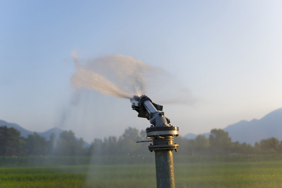 Close-up of an irrigation sprinkler spraying water on a field with mountains and trees in the background.