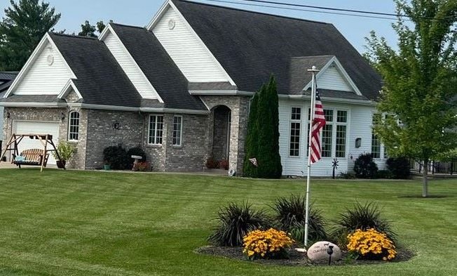 A charming two-story home with a blend of stone and white siding, featuring well-kept lawns and colorful flower beds with an American flag.