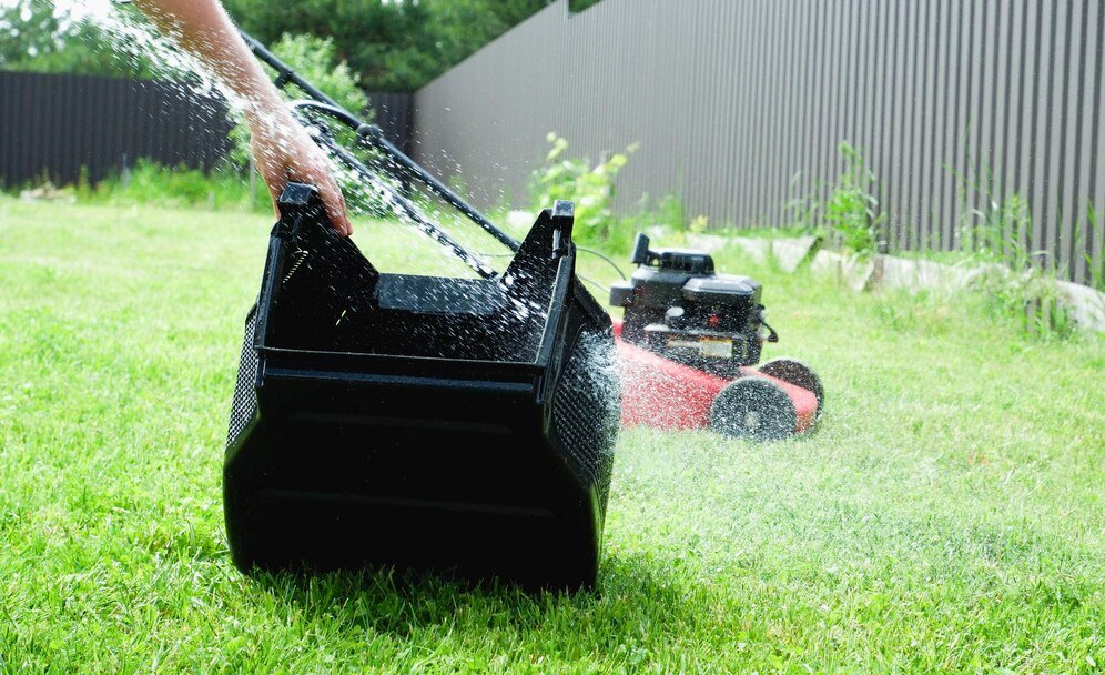 A person holds a grass catcher while cleaning a lawn mower, with grass clippings flying into the air against a green lawn backdrop.