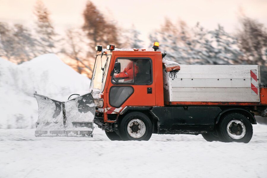 A snowplow truck clears snow from a snowy landscape, with a driver in bright orange attire at the controls.