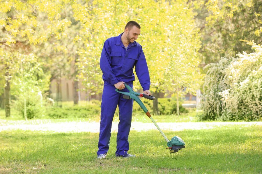 A person in a blue jumpsuit uses a trimmer to maintain a lush green lawn surrounded by vibrant trees and blooming flowers.