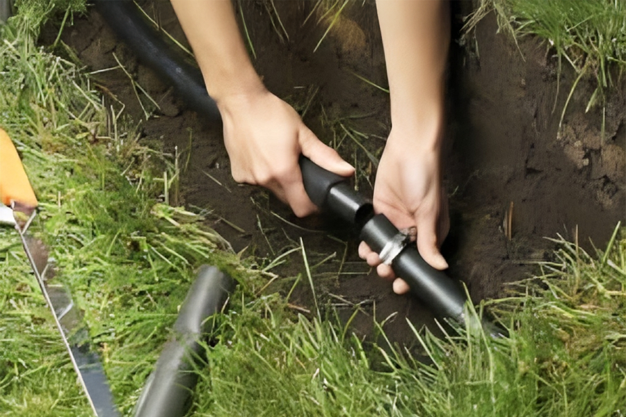 A person connects black irrigation tubing in a grassy area, using a tool to secure the joint in freshly dug soil.