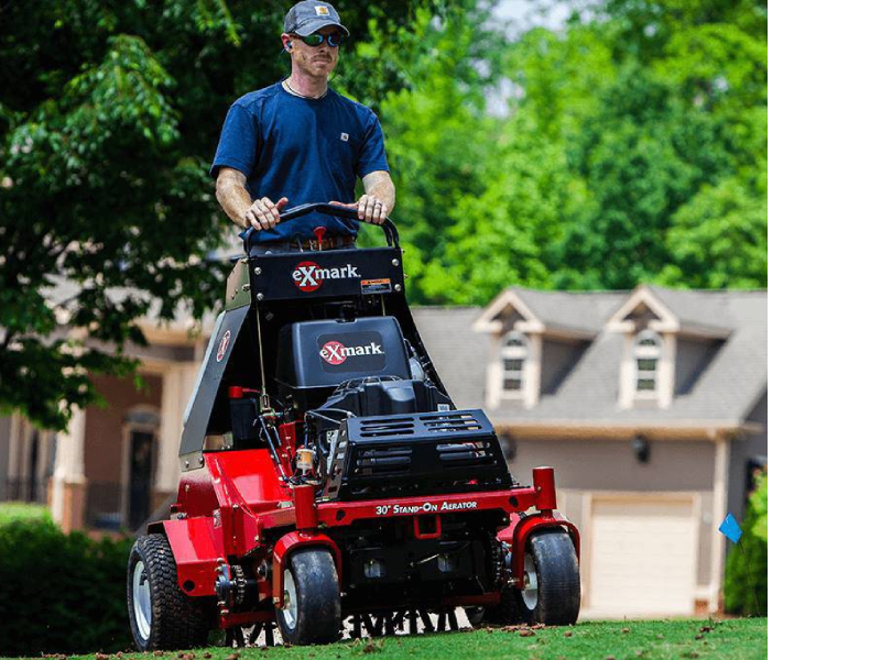 A person operates a red Exmark stand-on aerator on a lush green lawn, with a residential house visible in the background.